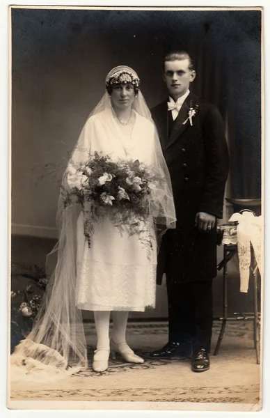 Vintage photo of newlyweds. Bride wears a long veil and holds wedding bouquet. Groom wears black suit and white bow tie. Black & white antique studio portrait.