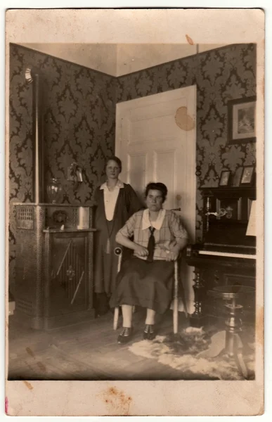 Vintage photo shows women pose at home. In the picture are piano and stove. Black & white antique photography.