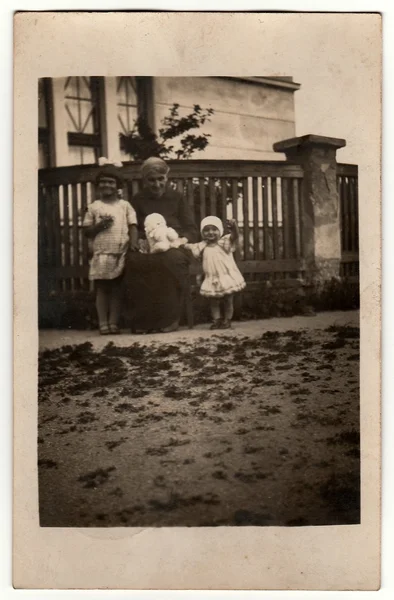 Vinatge photo shows woman and children pose in front of house. The woman holds teddy bear. Black & white antique photo.