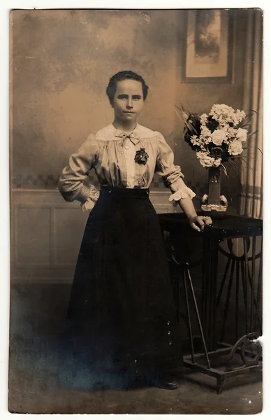Vintage photo shows woman wears an elegant dress, poses next to black table. On the table is vase with flowers. Black & white antique studio photography with sepia effect.