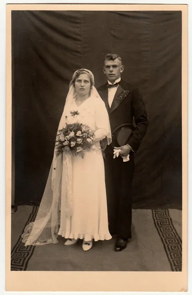 Vintage photo of newlyweds. Bride wears a long veil and holds wedding bouquet. Groom wears black suit and bow tie. Black & white antique studio portrait.