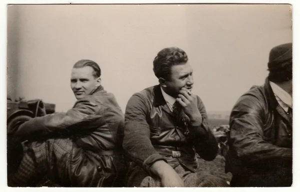 Vintage photo shows men wear leather jacket and vest. They have a rest after car trip. Black & white antique photography.