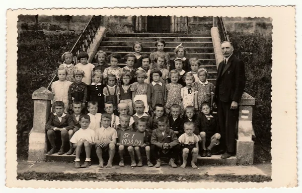 Vintage photo shows pupils (schoolmates) and their teacher pose in front of school. Black & white antique photography.