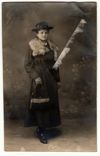 The vintage photo shows young woman wears an elegant coat with fur scarf, ladies wide-brimmed hat and holds handbag. The studio photography with sepia effect was taken in pre-twenties.
