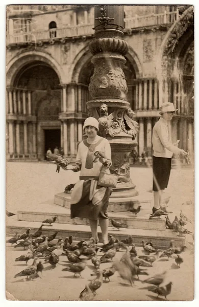 Vintage photo shows an elegant woman holds pigeons in hands. Second woman feeds pigeons/doves with grain. Women on holiday (vacation).