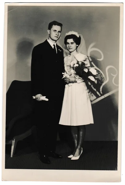 Retro photo shows bride with white kala flowers and groom wears a dark suit and white gloves. Black & white vintage photography