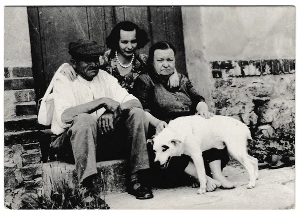 Retro photo shows rural people sit on a doorstep with dog. Black & white vintage photography
