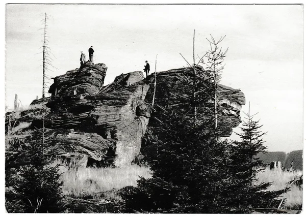 Retro photo shows tourists stand on top of the rock. People on holiday (vacation). The right side of rock remotely resamble snout of animal.  Black & white vintage photography