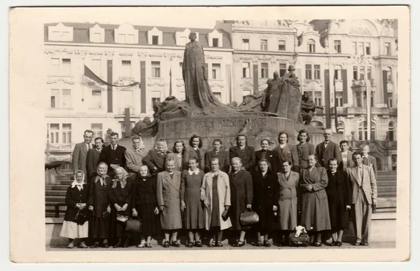 Vintage photo shows rural people pose in front of the Jan Hus Memorial at the Old Town Square. Retro black & white photography.