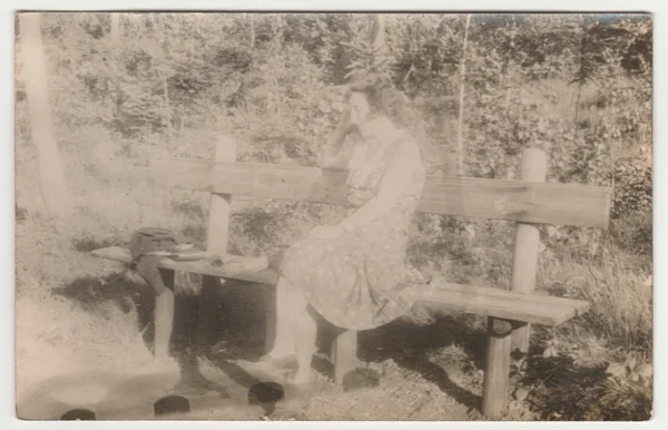 Vintage photo shows a young woman sits on the bench in the forest. Retro black & white photography. With original film grain, blur and scratches.
