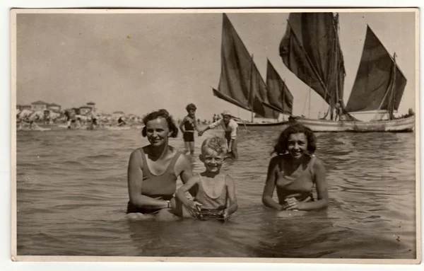 Vintage photo shows family (mother with son and daughter) have a swimm in the sea. Sailing boats are on the background. Vacation theme. Retro black & white photography.