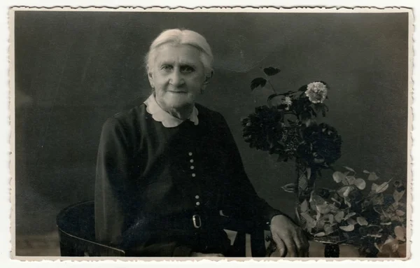 Vintage photo shows elderly woman sits on the period armchair in a photography studio. On the table are flowers. Retro black & white studio photography with sepia effect.