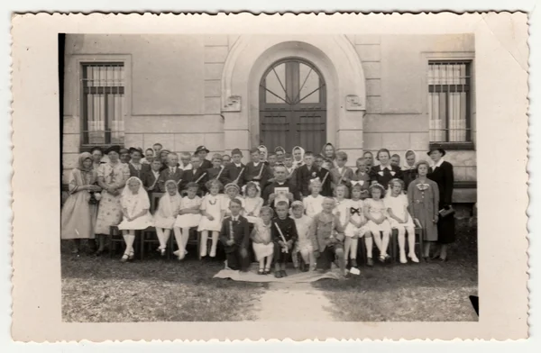 Vintage photo shows children and their relatives poses after a First Communion in front of the school. Retro black & white photography.