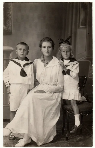 Vintage photo shows woman with her children (boy and girl). They wear sailor costumes. Retro black & white studio photography.