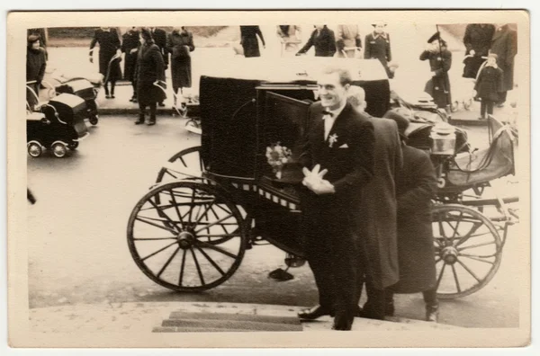 Vintage photo shows a groom. A historical carriage (coach)  is on the background and historical prams too. Retro black & white photography.