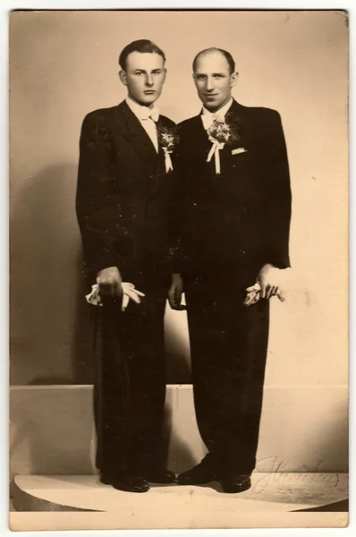 Vintage photo shows two men (groom and his best man) pose in photography studio. Retro black & white photography.