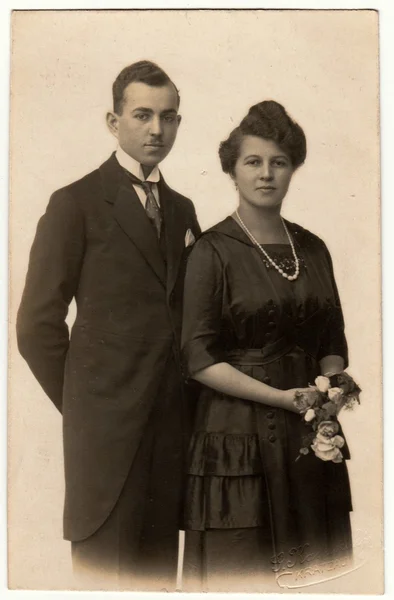 Vintage photo shows a young couple wears black clothes. Woman holds flowers. Retro black & white photography.