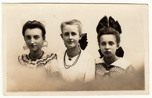Vintage photo shows a group of girls poses in the photography studio. Girls wears hair ribbon and some of them has necklace ((string of) beads ). Retro black & white photography with sepia effect.