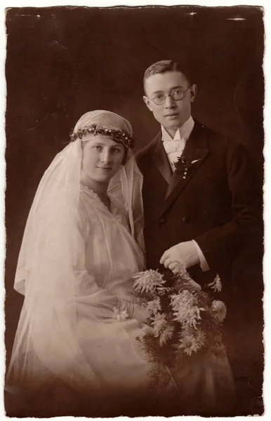 Vintage photo shows newlyweds. Groom wears glasses and bride wears long veil. Retro black & white wedding photography.