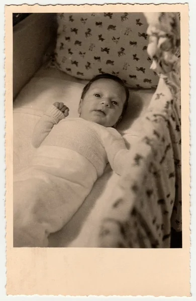 Vintage photo shows cute small baby in a cot (crib, baby bed). Retro black & white photography.