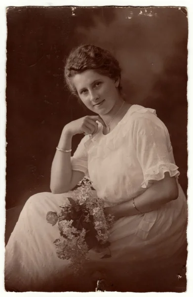 Vintage photo shows woman with short hair. Woman wears white dress and holds bouquet (bunch of flowers). Retro black & white studio photography with sepia effect.