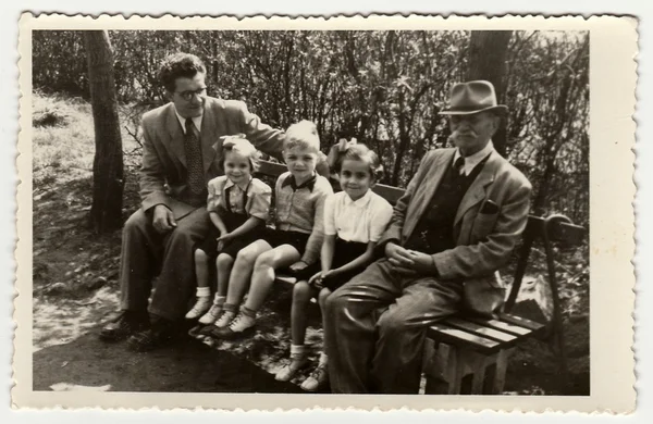 A vintage photo shows men with children. They sit on a bench, circa 1940.