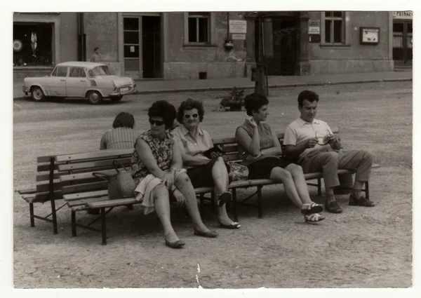 Vintage photo shows people sit on a bench, circa 1950s.