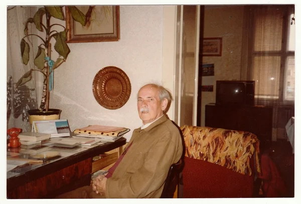 Vintage photo shows man sits on chair, circa 1980s.