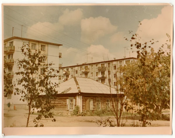 Vintage photo shows log cabin (log house) and blocks of flats in the background.