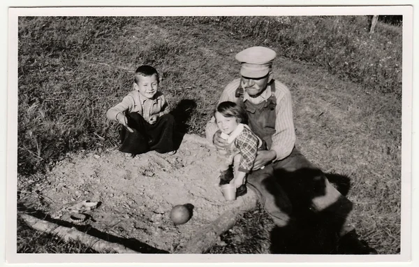 A vintage photo shows grandfather plays with children on sandpit. Antique black & white photo shows photographer shadow.