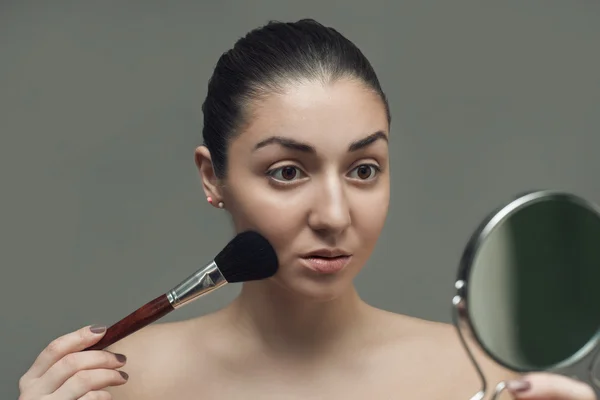 Beautiful girl looking in the mirror and applying cosmetic with a big brush.