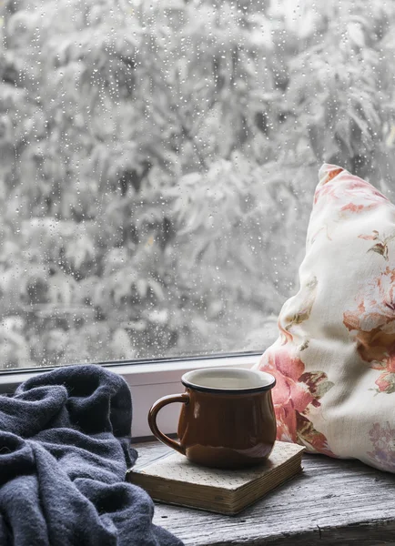Coffee mug, book, pillows and a plaid on the light wooden surface against window with rainy day view. Vintage style. The concept of homeliness and comfort
