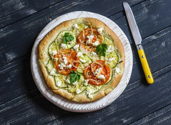 Pizza with zucchini, tomatoes, onions and feta cheese on a light board on dark wooden background. Delicious food