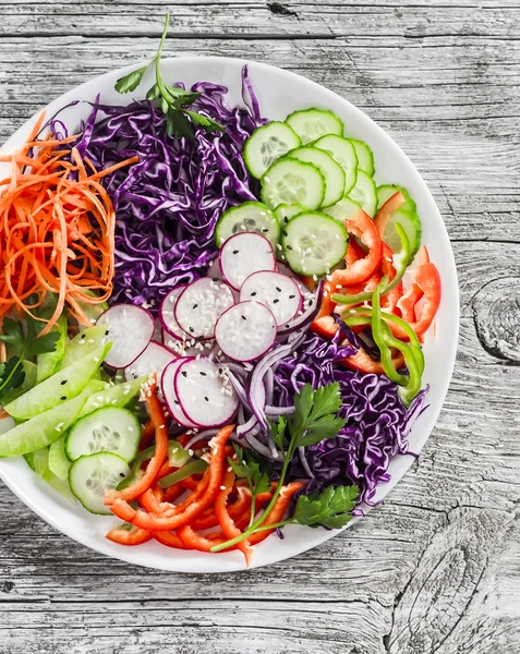 Fresh vegetable salad with red cabbage, cucumber, radish, carrots, sweet peppers, red onion and parsley on a white plate.  On wooden rustic background