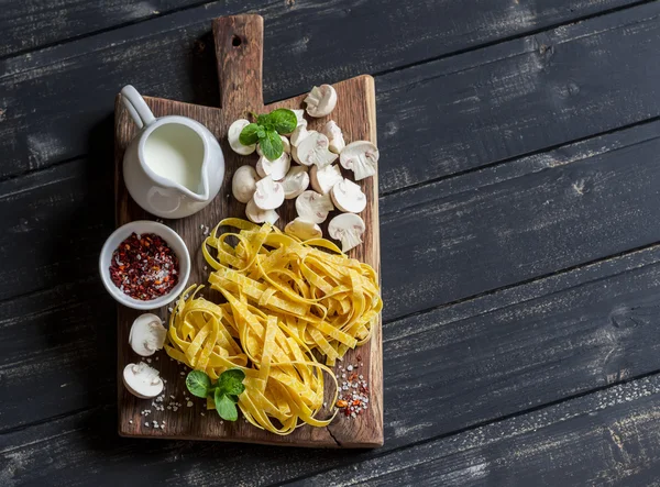 Raw ingredients for cooking pasta with mushroom cream sauce - pasta, mushrooms, cream, spices. On rustic wooden board on dark background