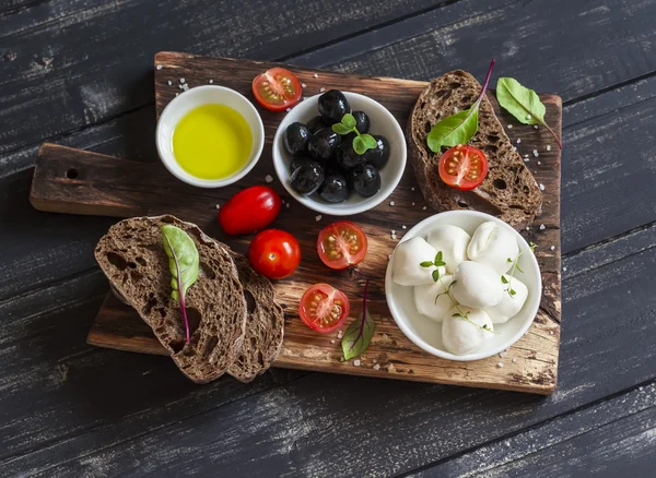 Mediterranean snack - mozzarella, olives, rye ciabatta bread, cherry tomatoes on a rustic wooden cutting board on a dark background