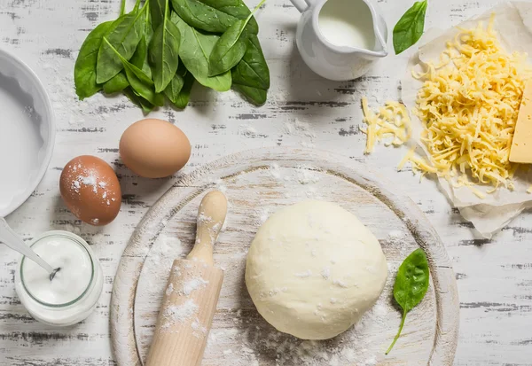 Dough, eggs, fresh spinach, cheese - raw ingredients to prepare pie with spinach, cheese and egg. On a light wooden background. Baking background
