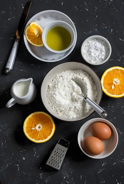 Ingredients for making orange cake with olive oil - flour, eggs, olive oil, powdered sugar on a dark stone background