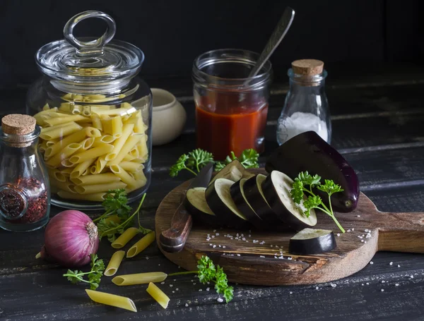 Penne pasta, eggplant, onion, tomato sauce, spices and herbs - raw ingredients for cooking Italian pasta with eggplant . On a dark wooden background. Healthy vegetarian food