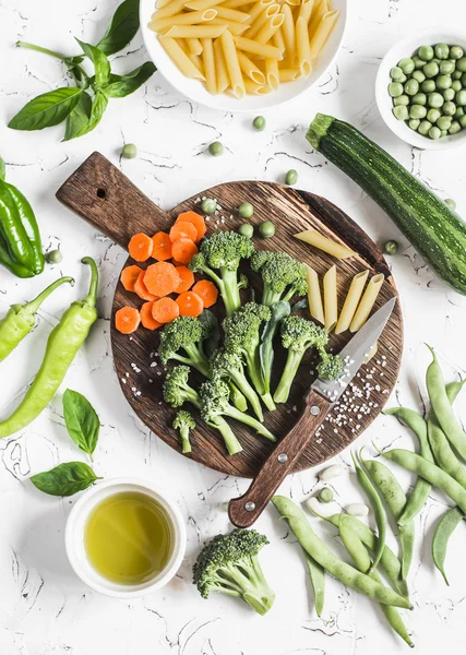 Raw ingredients for cooking vegetarian lunch - dry pasta, fresh broccoli, zucchini, green peas and beans, carrots, olive oil. On a light background, top view