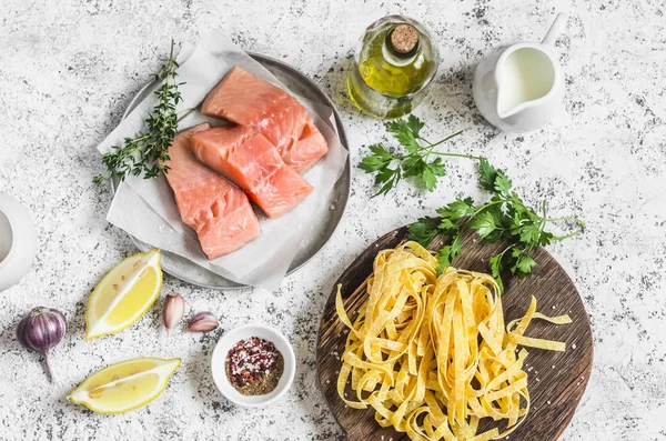 Ingredients for cooking lunch - raw salmon, dry pasta tagliatelle, cream, olive oil, spices and herbs. On a light background, top view