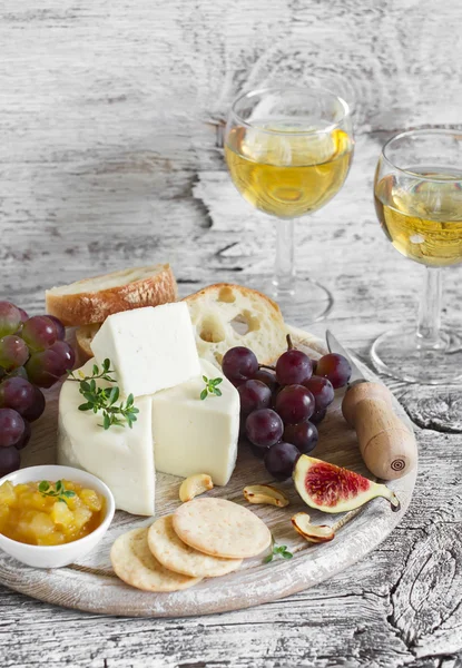 Delicious appetizer to wine - ham, cheese, grapes, crackers, figs, nuts, jam, served on a light wooden board, and two glasses with white wine