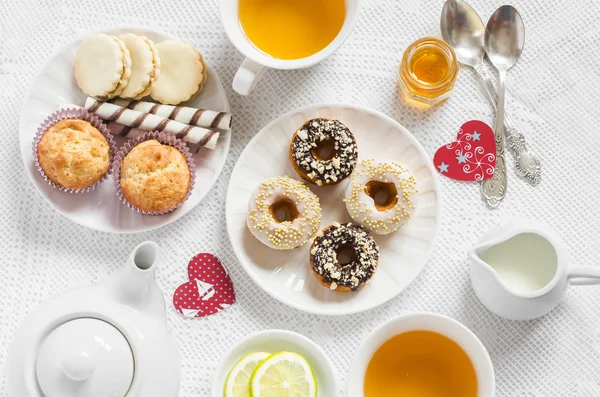 Valentine\'s day romantic Breakfast. Lemon green tea and sweets - banana muffins, cookies with caramel and nuts, donuts with chocolate and lemon glaze, tea set, red paper hearts on a white tablecloth on a light surface.  Tea time.