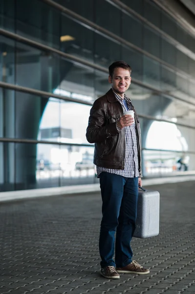 Stylish handsome young male traveller with bristle standing outdoors near the airport terminal. Man wearing jacket and shirt. Smiling person looking to camera holding cup of coffee