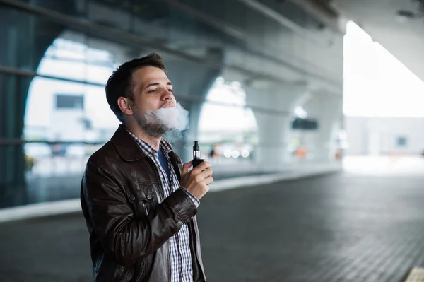 Young man enjoying a satisfying e-cigarette standing in profile against airport terminal background