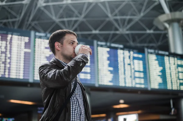 Young man with a bag in airport near flight timetable drinking coffee