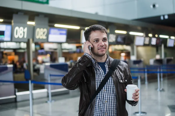 Portrait of young handsome man walking in modern airport terminal, talking smart phone, travelling with bag, wearing casual style clothes, blurred registration desks on background