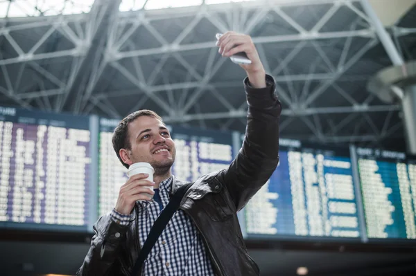 Smiling young traveller man with a coffee at the airport makes selfie in front of timetable board
