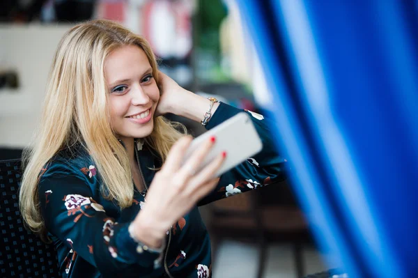 Gorgeous smiling blonde woman making self portrait with mobile phone camera while sitting in modern cafe inside, charming happy female posing while photographing herself for social network picture