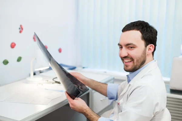 Young caucasian man doctor examines MRI image of human head in office looking at the patient and smiling